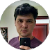 Uma pessoa com cabelo curto e escuro, vestindo uma camisa roxa, está tirando uma selfie no espelho com um smartphone. O plano de fundo mostra parte de uma porta vermelha e uma parede com algumas decorações com o tema Cloudflare.